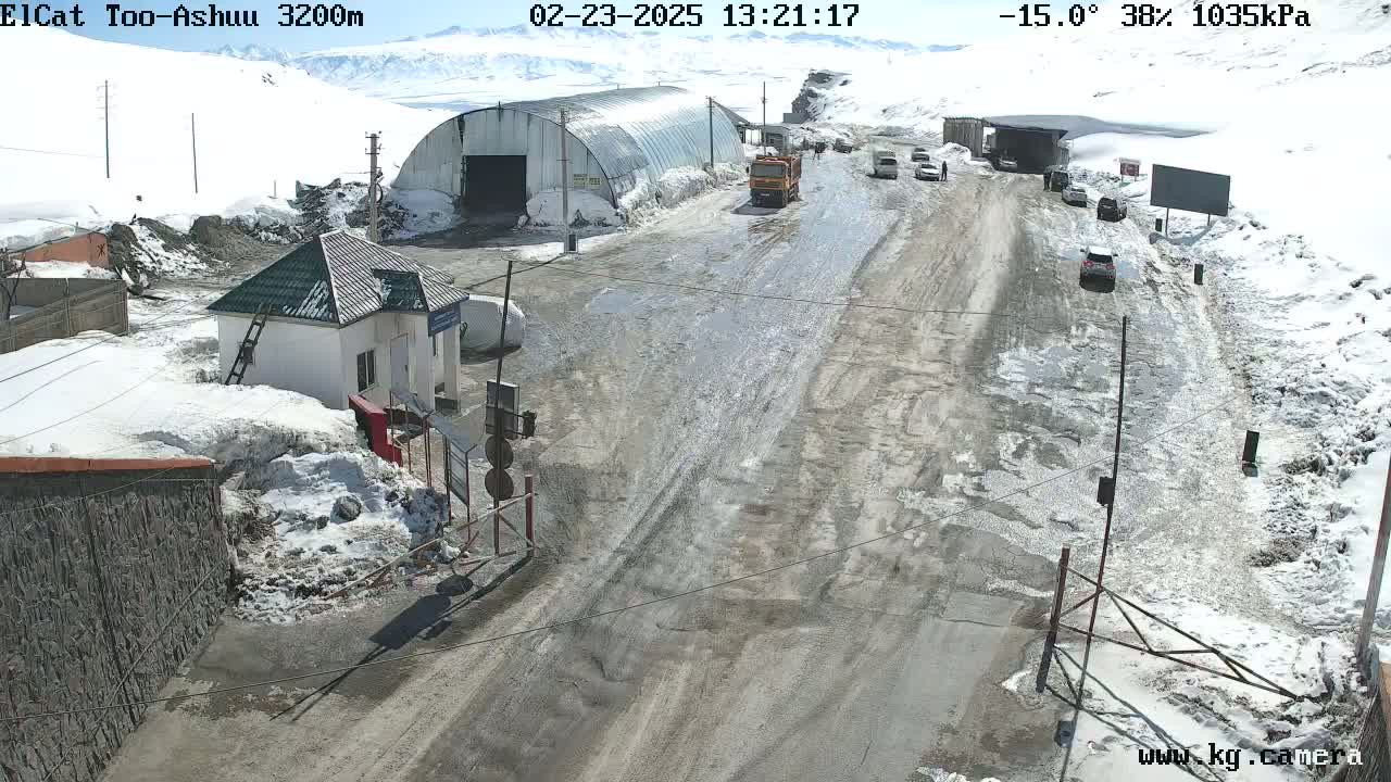 A wet, snow-covered road with several vehicles and a small building is seen on a sunny, cold day.