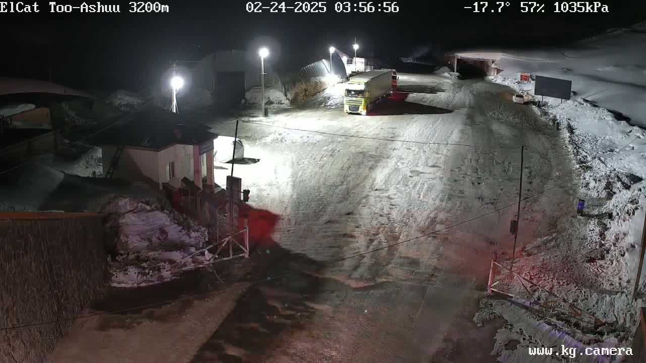 A snow-covered road at night with a few vehicles, including a semi-truck, near a small building and a gate under bright lights.