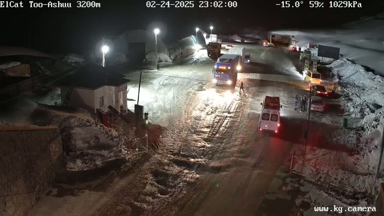 A snow-covered road at night with several vehicles, including trucks and cars, and a few people near a small building.