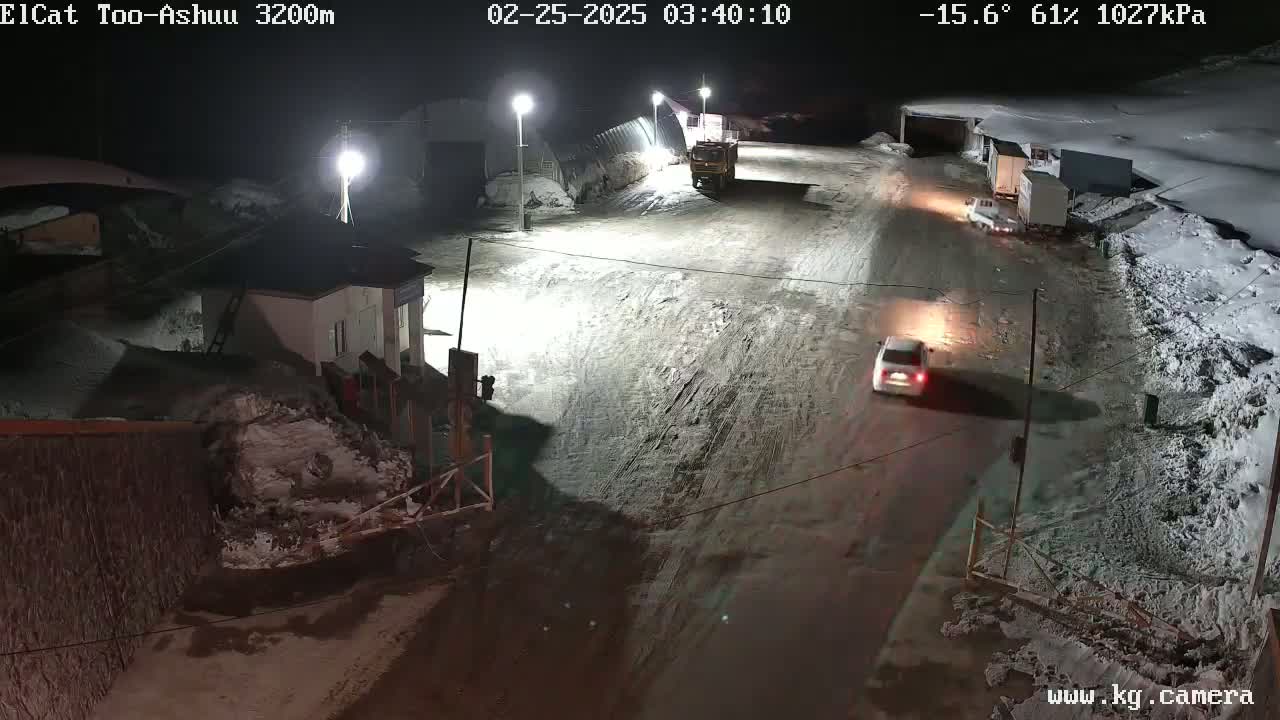A snow-covered road at night, illuminated by streetlights, shows several vehicles parked and driving near buildings.