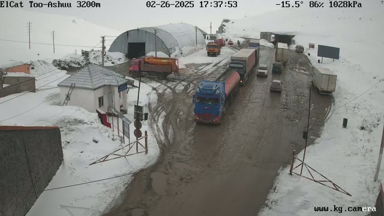 A muddy road with several trucks and cars, surrounded by snow-covered ground and buildings in cold, snowy weather.