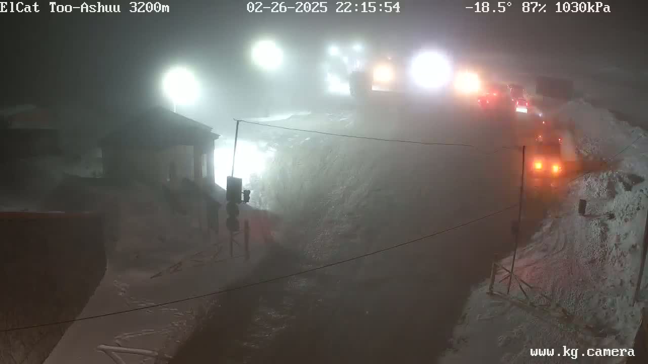 A nighttime, snowy scene shows a fog-shrouded road with vehicles and snowplows, lit by artificial lights.