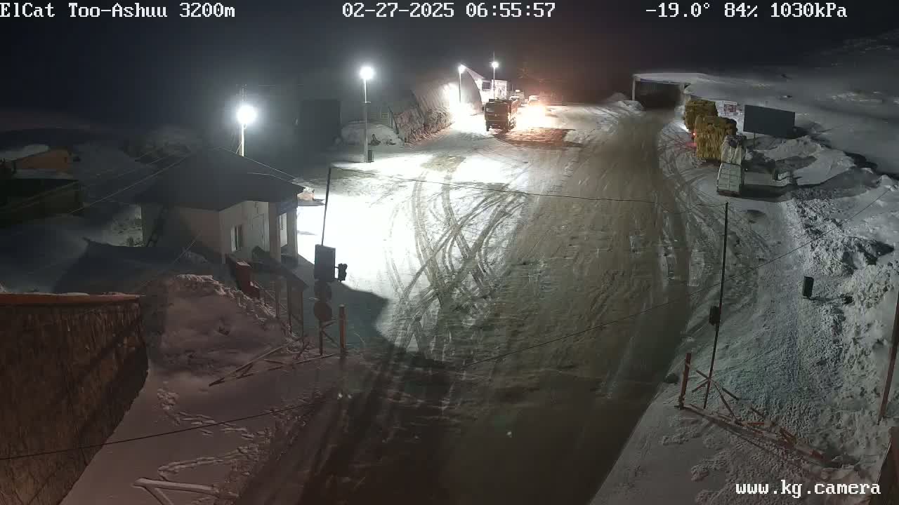 A snow-covered road at night with several vehicles and buildings visible under bright lights.
