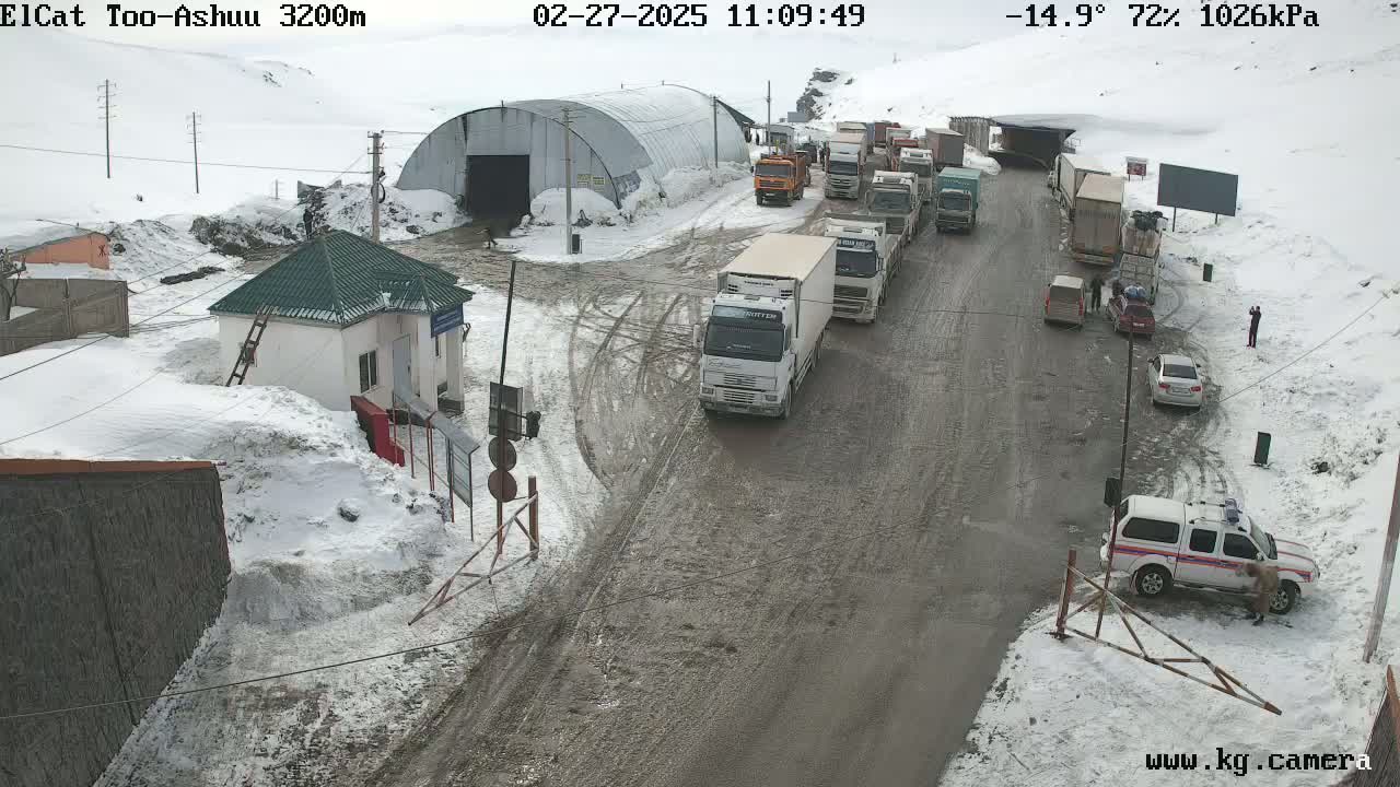 A snowy, muddy roadway shows a line of trucks and cars at a border crossing, with a small building and snow-covered landscape surrounding it.