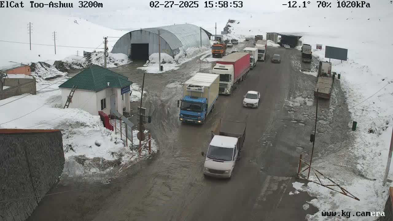 A snowy, muddy road with several trucks and cars, including a line of trucks stopped near a building and a smaller structure, in what appears to be a mountain pass.