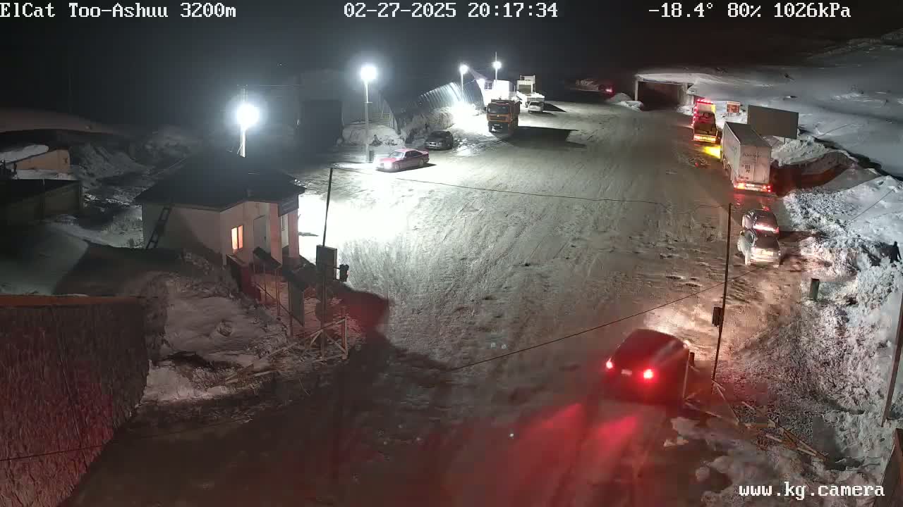 A nighttime, snowy scene shows several vehicles on a wet road near a small building, with snow banks lining the sides of the road.