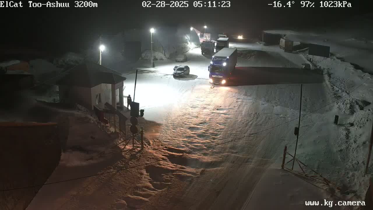 A snow-covered road at night is illuminated by streetlights, with several vehicles, including a large truck, visible near a small building.