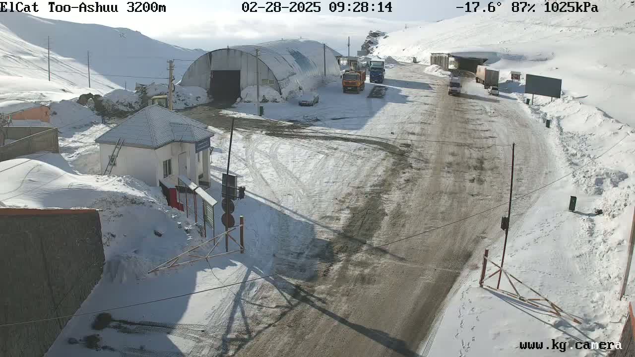 A snow-covered mountain road scene with several vehicles near a building and tunnel, under a sunny sky.