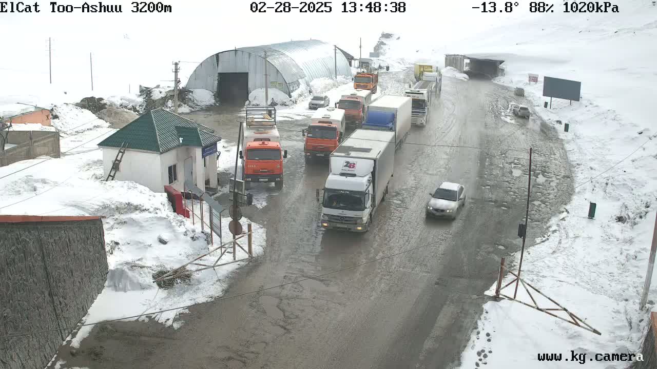 A snowy, wet road with several trucks and cars, some stopped near a small building and a larger structure.