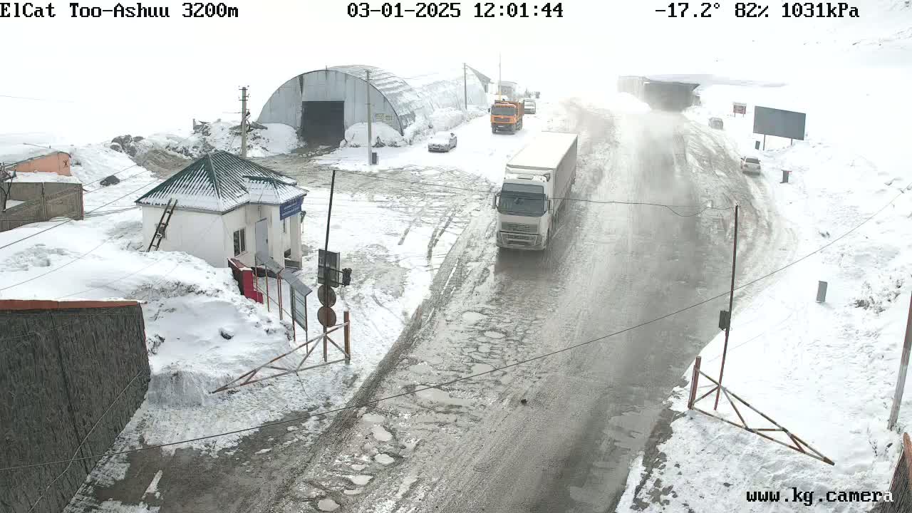 A snow-covered road with several vehicles, including trucks and cars, is seen near a small building and a large metal structure in a snowy, overcast landscape.