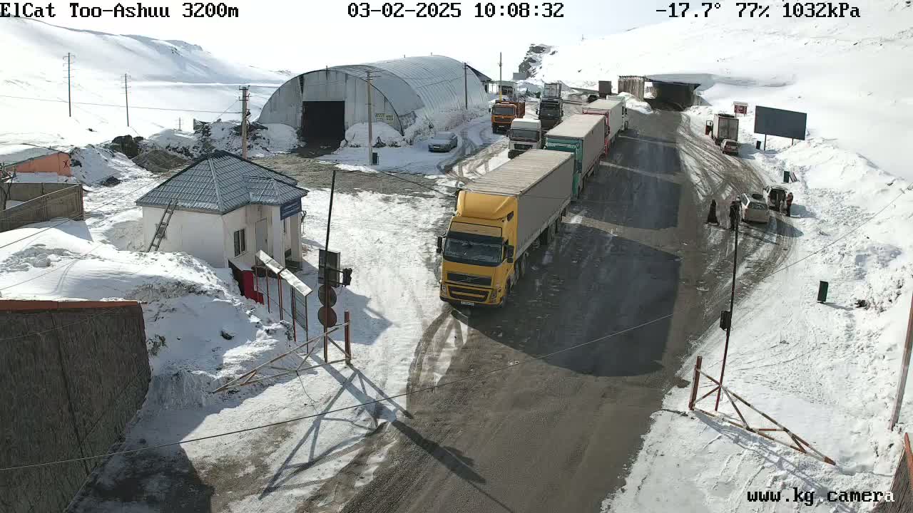 A line of trucks and cars are stopped at a snowy mountain pass checkpoint under a sunny sky.