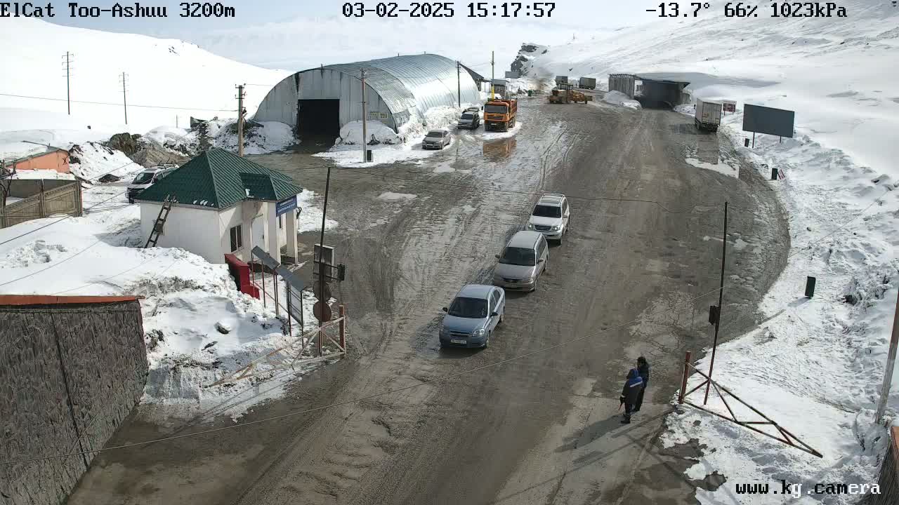 A muddy road in a snowy, mountainous area has several cars and trucks parked near a small building and a large metal structure; a person is walking near a gate.