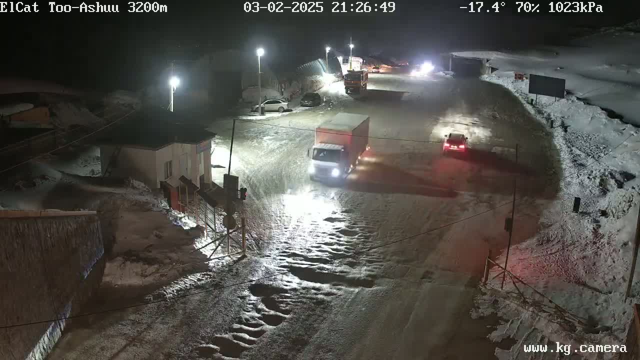 A snow-covered road at night with several vehicles, including a truck and cars, is illuminated by streetlights.