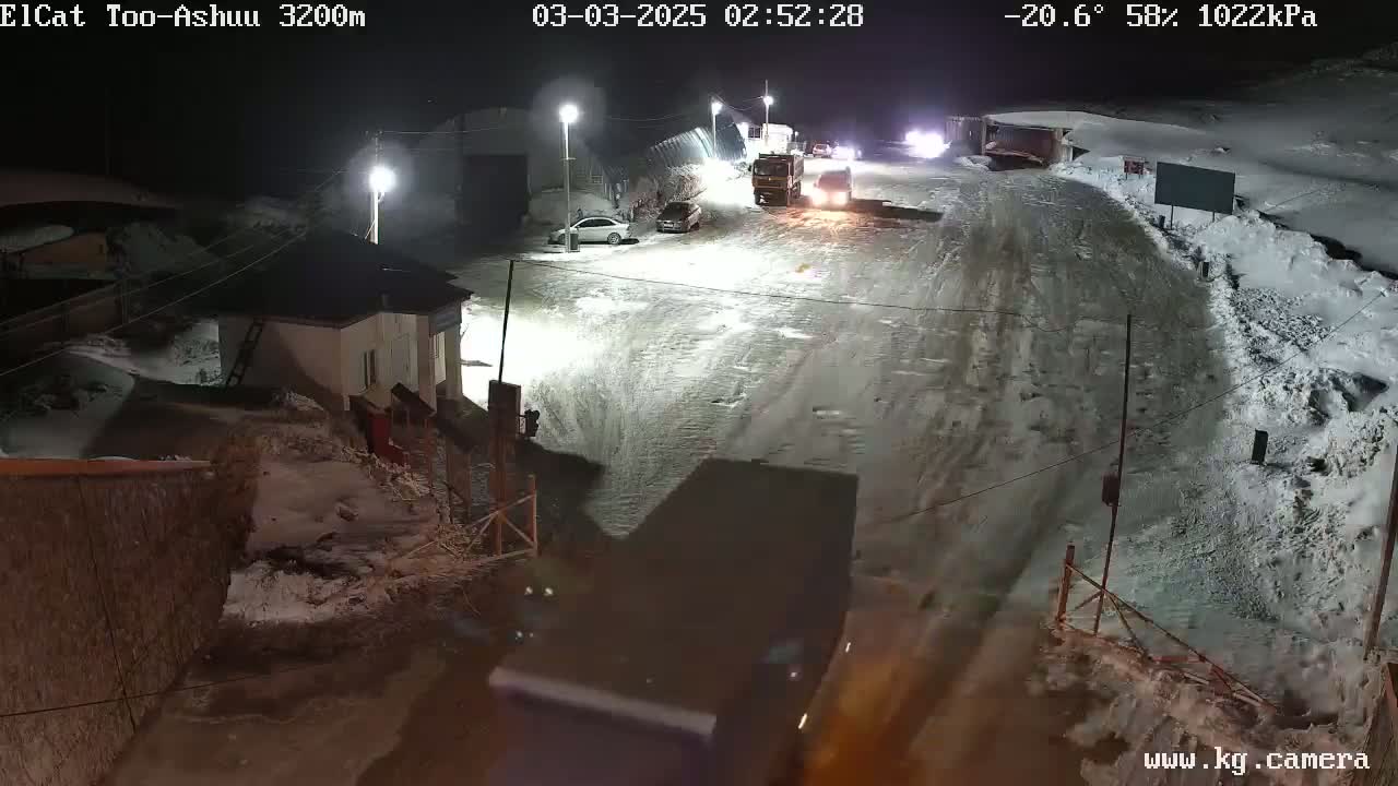 A snow-covered road at night, illuminated by streetlights, shows several vehicles including trucks and cars, near a small building.