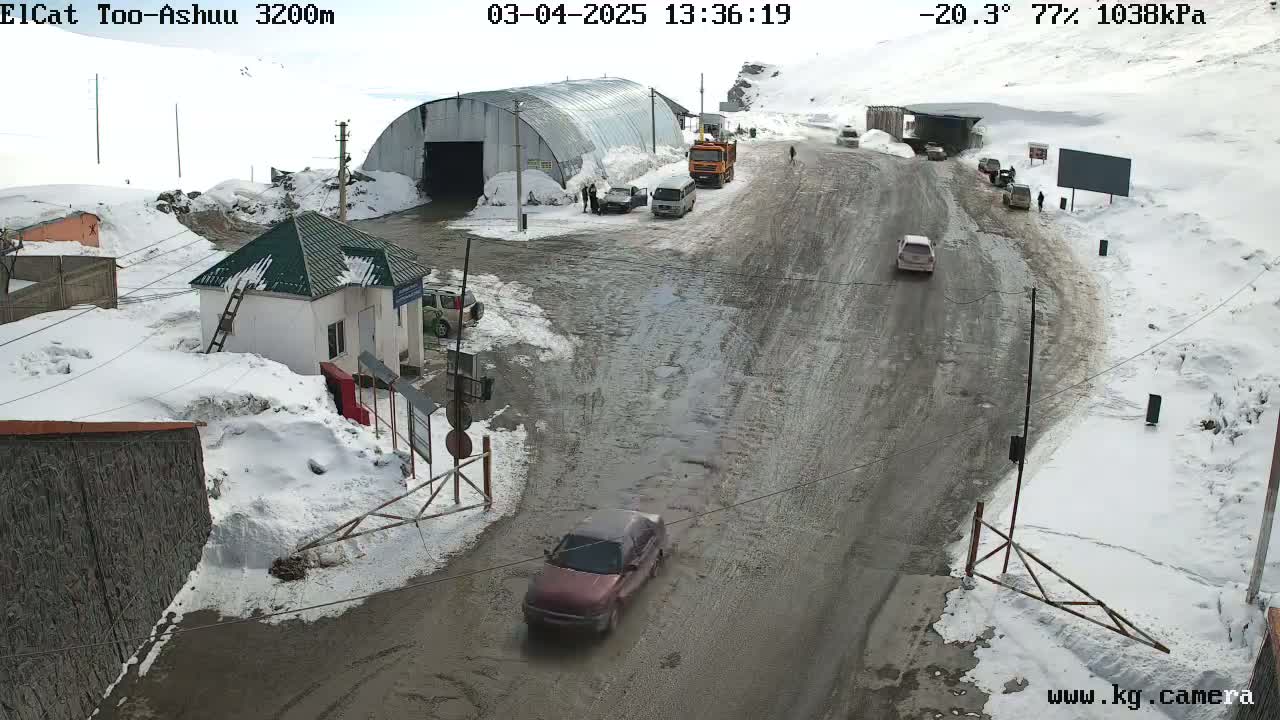 A snowy mountain road with several vehicles, a small building, and a large metal structure, under a cloudy sky with a temperature of -20.3°C.