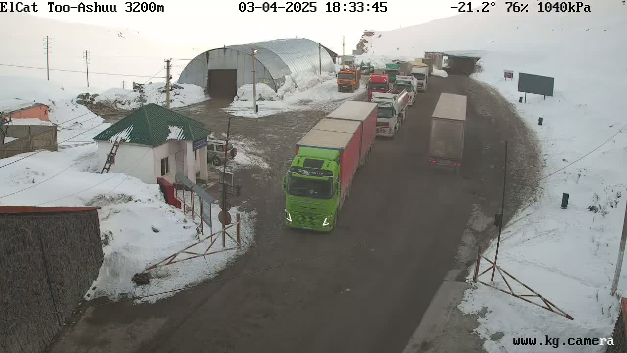 A line of trucks and other vehicles are navigating a snowy roadway near a building and a small guardhouse.