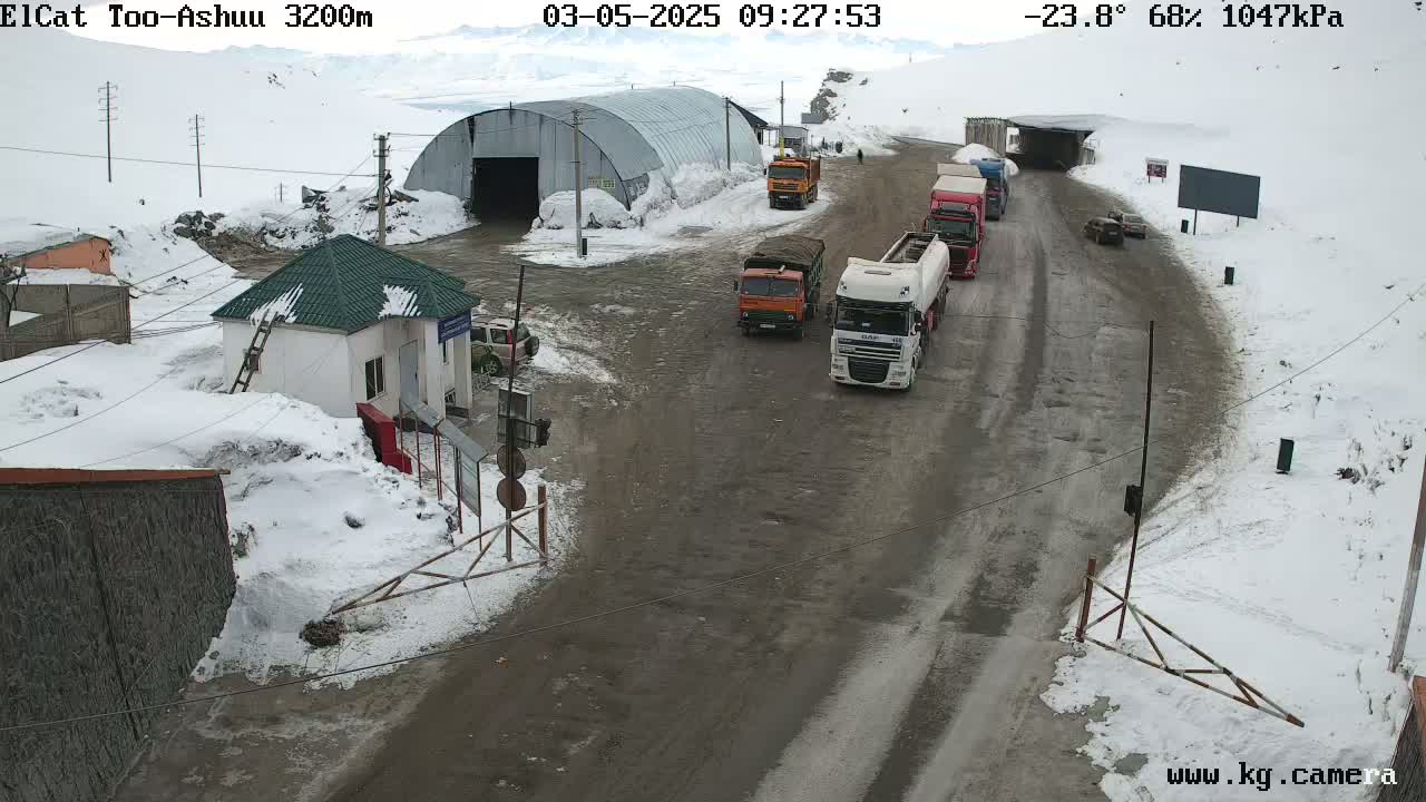 A snowy mountain road with several trucks and cars near a small building and a large metal structure, under a cloudy sky.