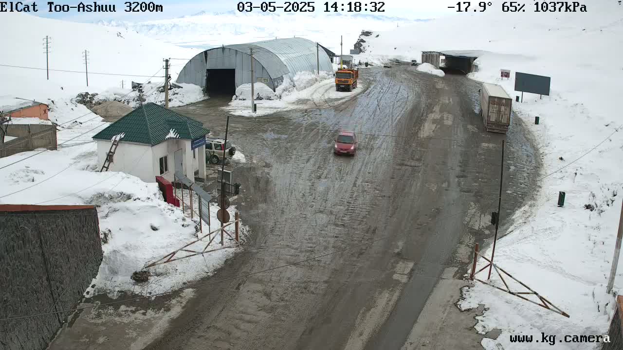 A muddy road winds through a snowy mountain pass, where several vehicles are visible near a small building and a large metal structure.
