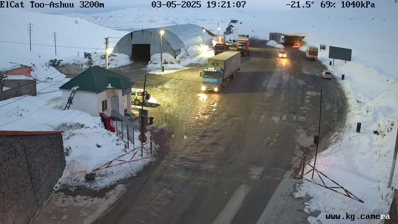 Several vehicles, including trucks and cars, are traveling on a wet, snow-covered road near a building and a checkpoint in a cold, nighttime environment.