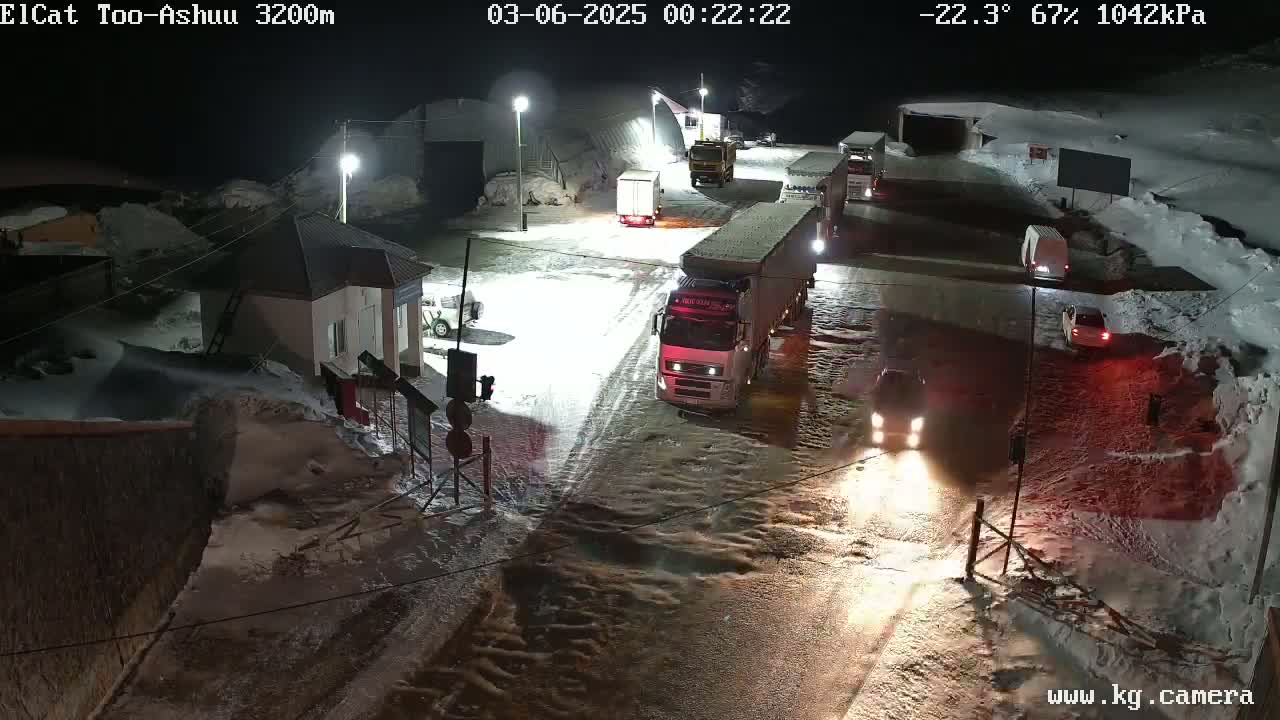 Several vehicles are parked and driving on a snow-covered road at night, near a small building with lights on.
