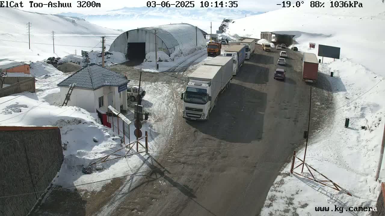 A snowy mountain pass road shows several vehicles, including trucks and cars, lined up near a small building and a large metal structure, under a partly sunny sky.