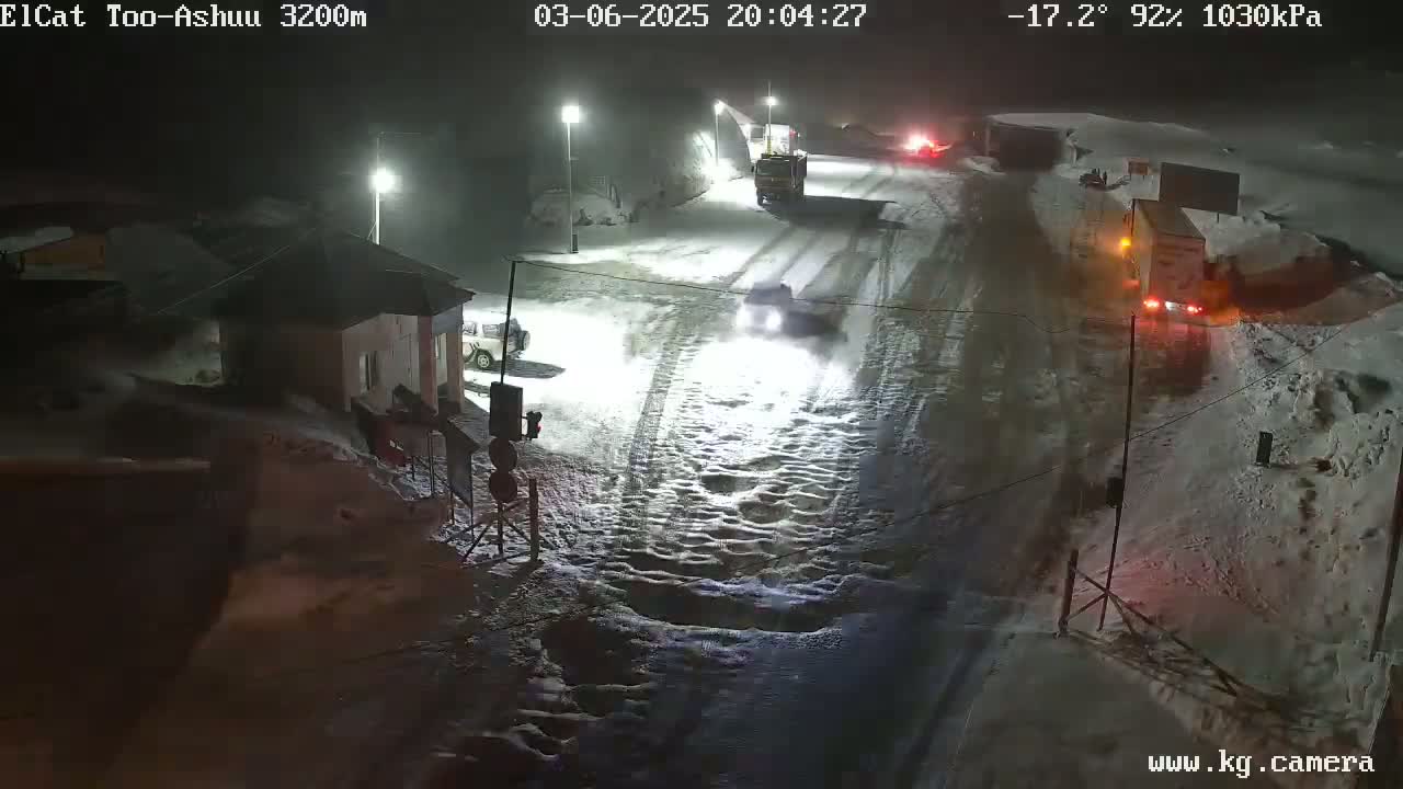 A snow-covered road at night with several vehicles, including trucks, driving through a dimly lit area near a small building.