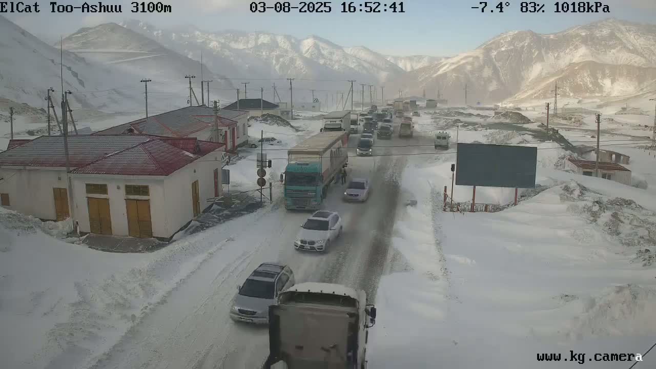 A snow-covered road with a line of vehicles, including cars and trucks, passing through a snowy mountain pass near some buildings.