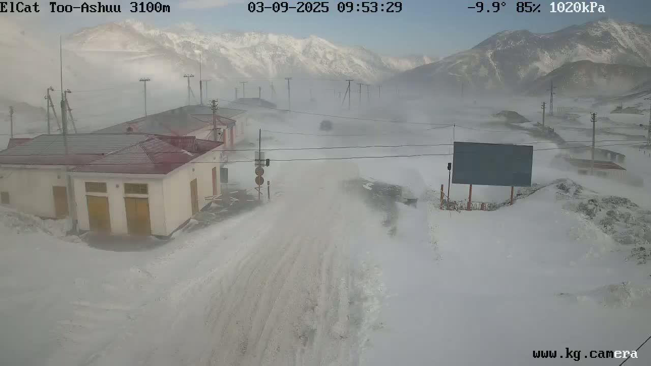 A snow-covered road and buildings are seen in a blizzard, with mountains in the background.