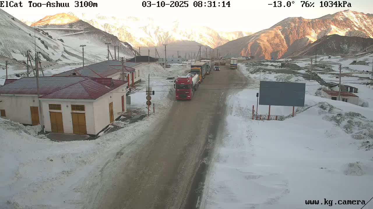 A snow-covered mountain pass road with several trucks and buildings, under a partly sunny sky with a temperature of -13 degrees Celsius.