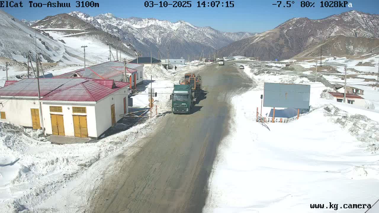 A mountain road scene shows several buildings and trucks on a snowy, sunny day.