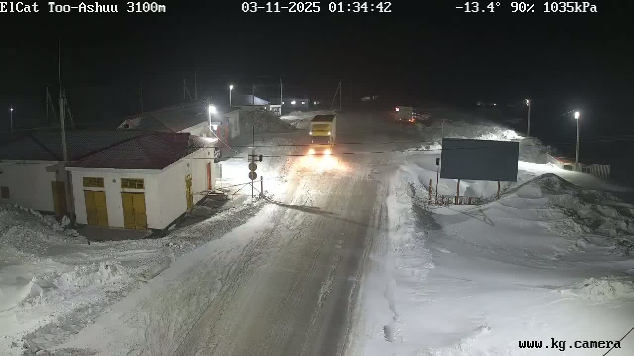 A truck drives down a snow-covered road at night, passing buildings under nighttime illumination.