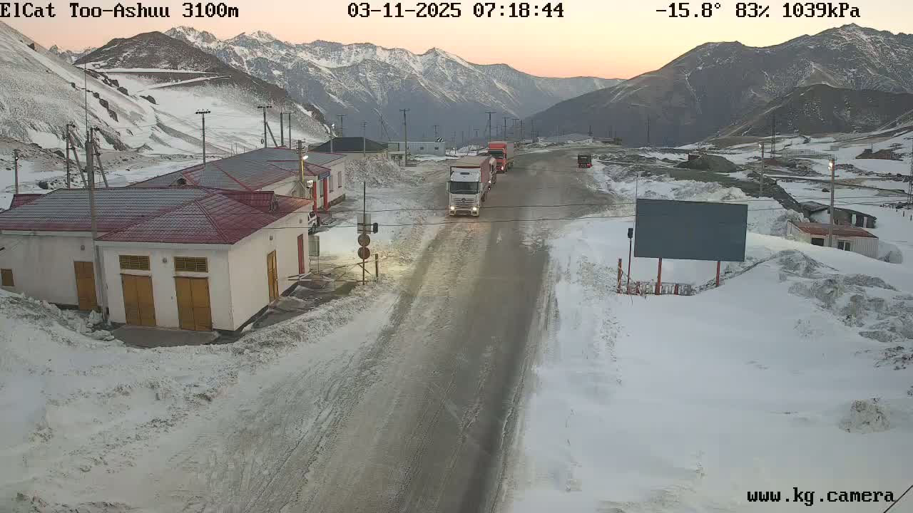 A snow-covered mountain road with several trucks and buildings, under a twilight sky.
