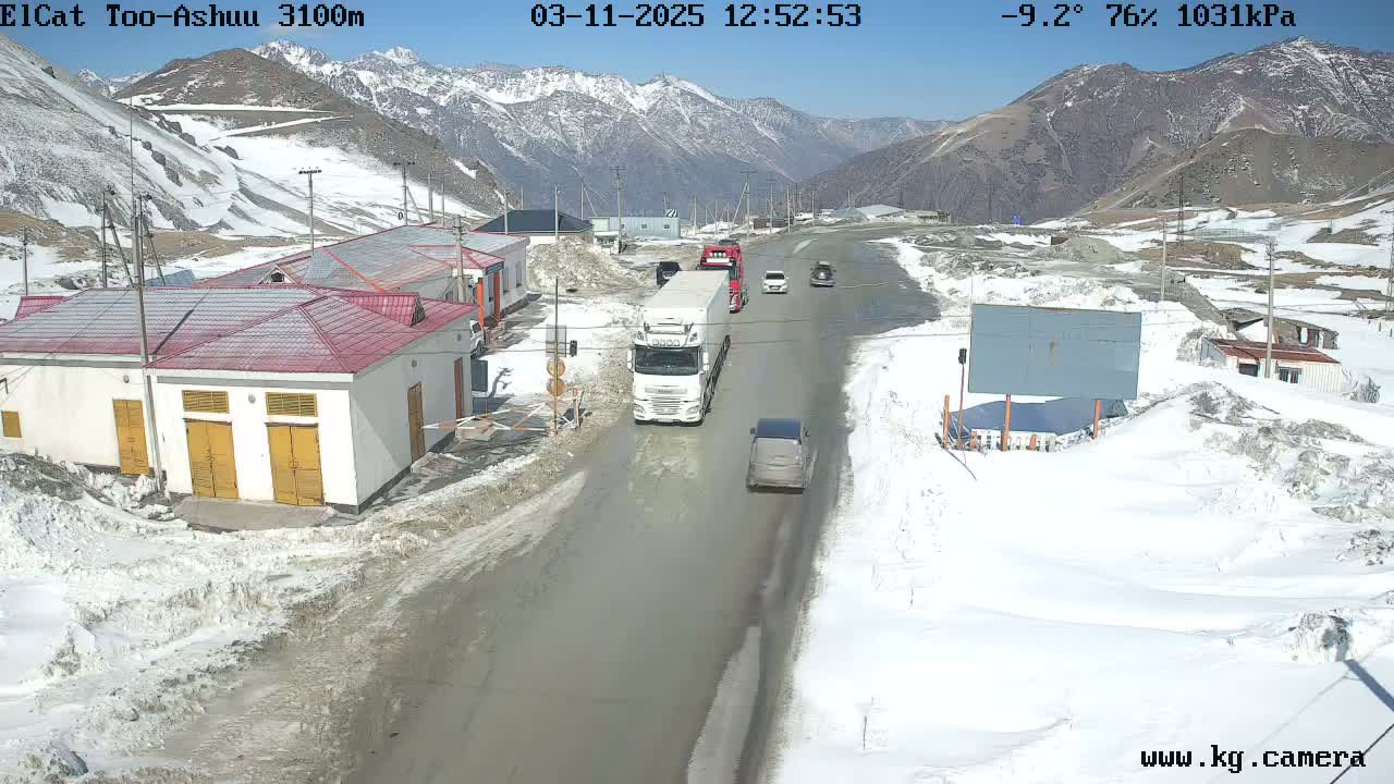 A mountain road scene on a sunny day with snow on the ground and mountains in the background shows several vehicles, including trucks and cars, traveling along a road passing by buildings.