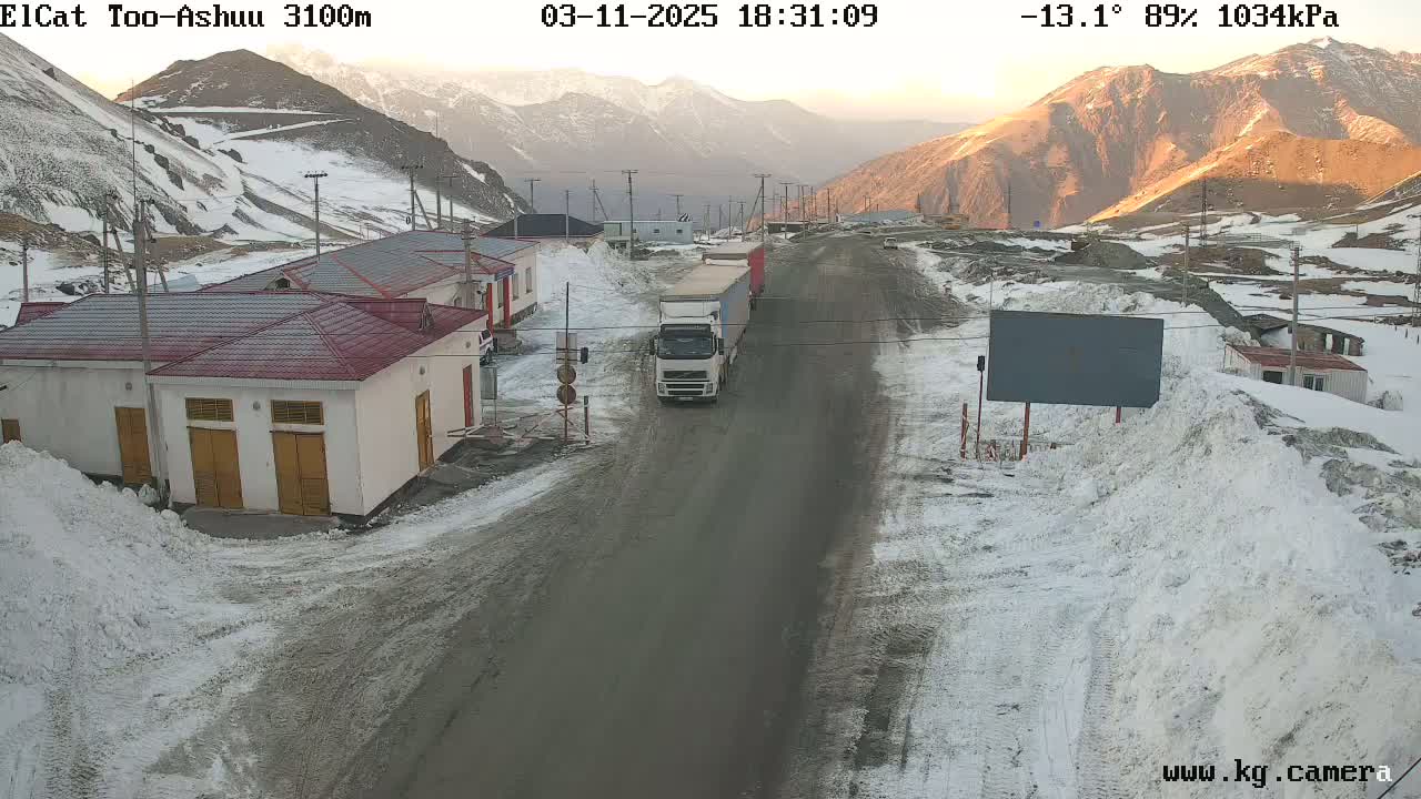 A snowy mountain road with buildings and a truck, under a partly sunny sky.