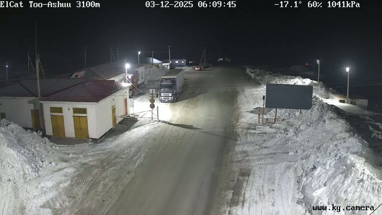 A nighttime, snowy scene shows a road with a truck parked near several buildings, with snow piled along the sides of the road.