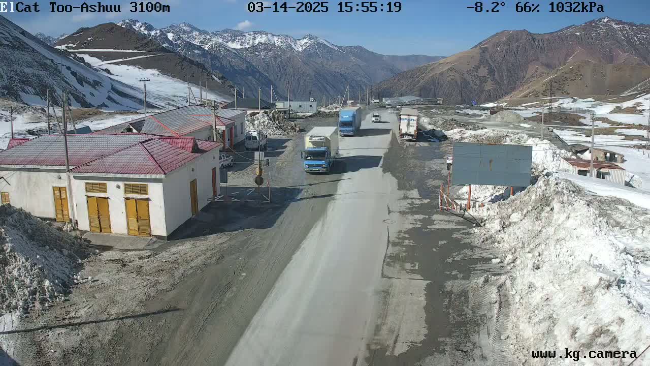 A mountain road with several trucks and a building complex, under sunny skies with patches of snow and ice on the ground and surrounding mountains.