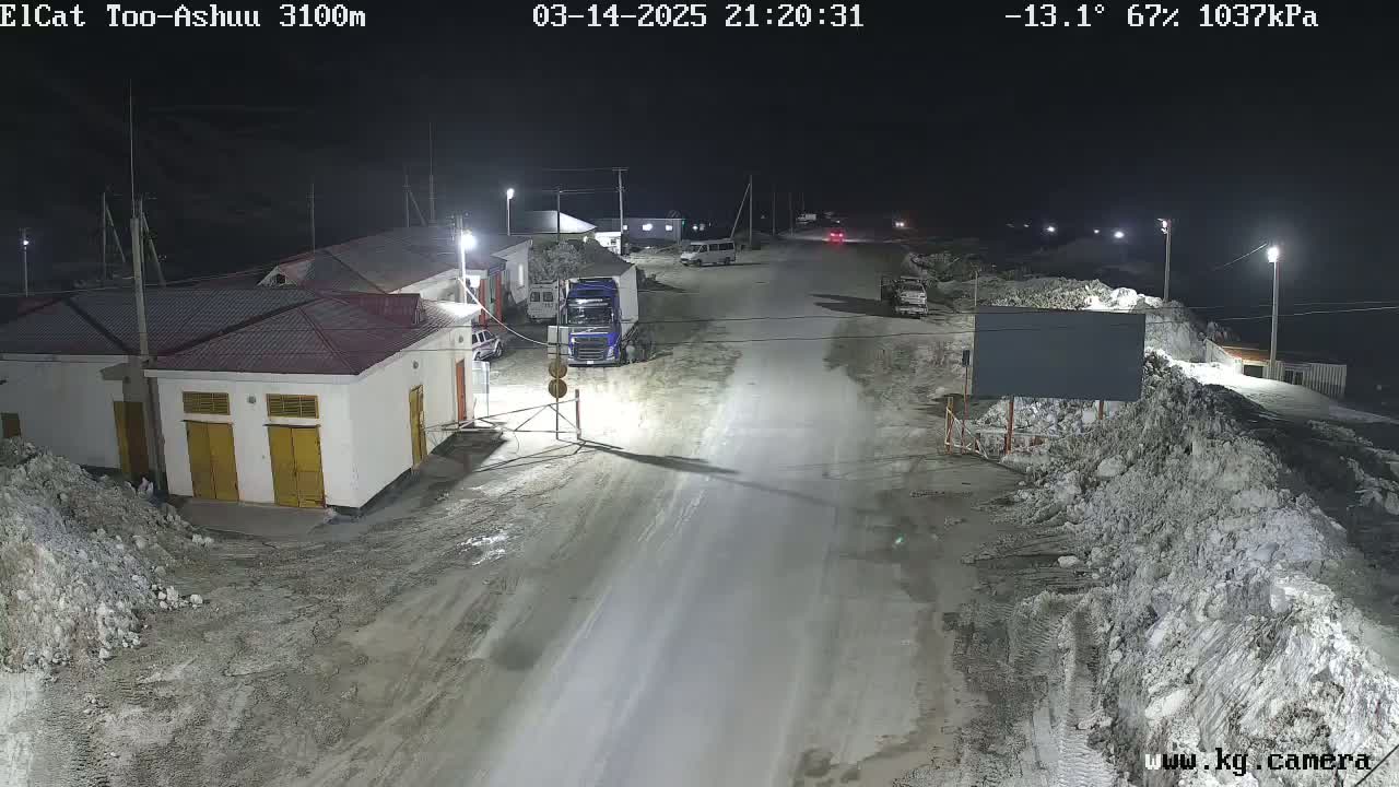 A nighttime, snow-covered road scene shows several vehicles parked near a building complex under artificial light, with a temperature of -13.1°C and 67% humidity.