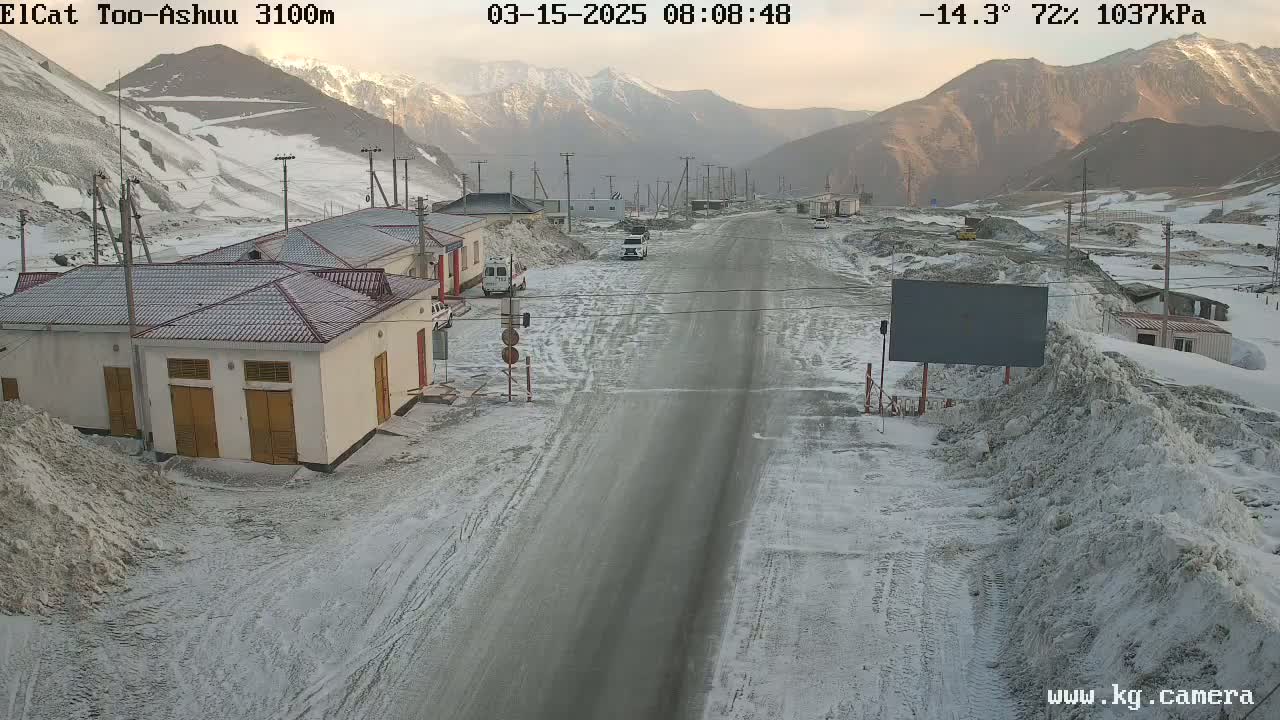 A snow-covered road winds through a mountainous valley, past small buildings and piles of snow, under a mostly clear sky.