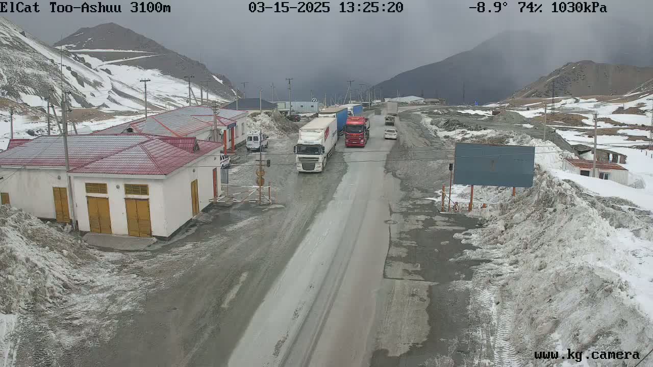 A snowy mountain pass road with several trucks and cars, some buildings, and snow banks lining the sides of the road under an overcast sky.
