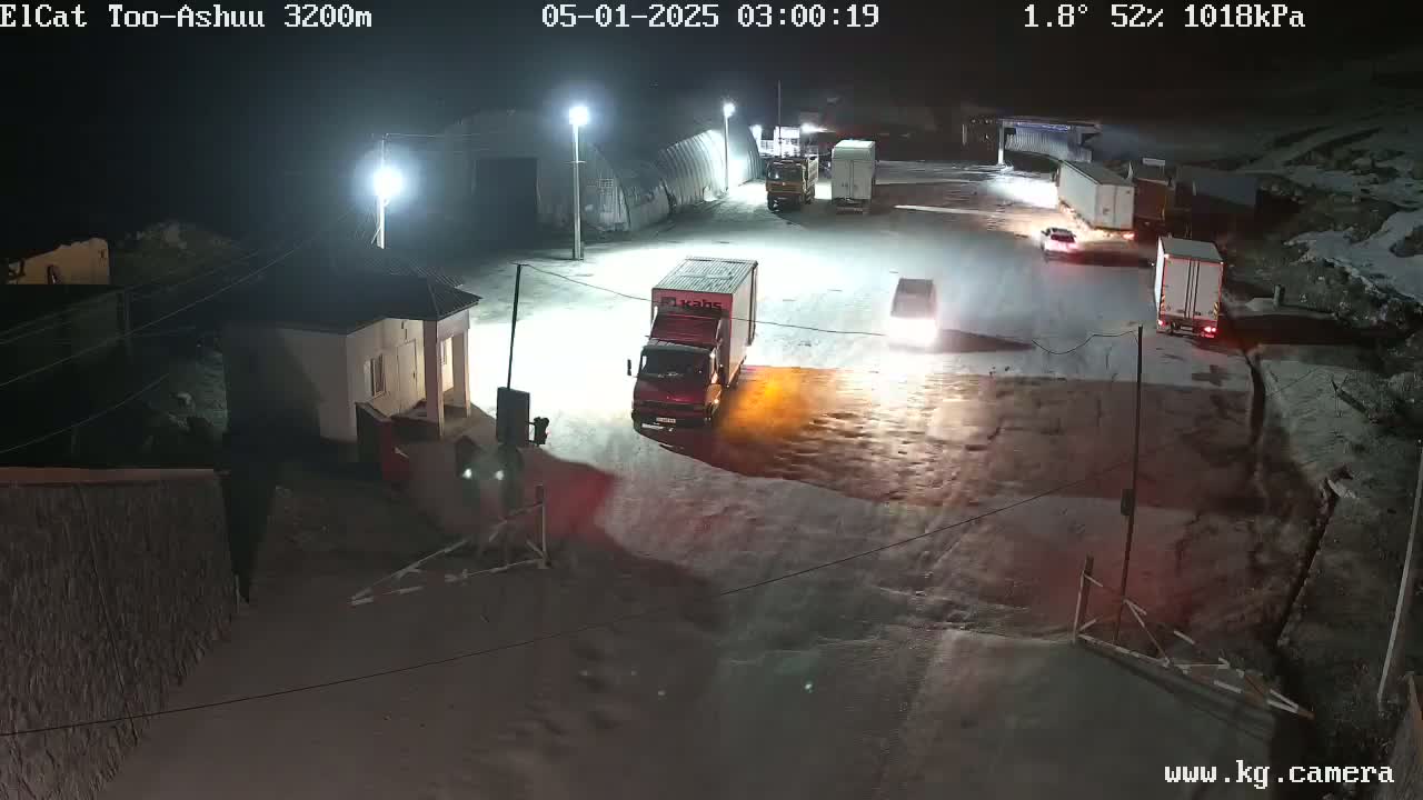 At night in snowy conditions, several trucks and cars are parked or moving slowly in a dimly lit area near a building.