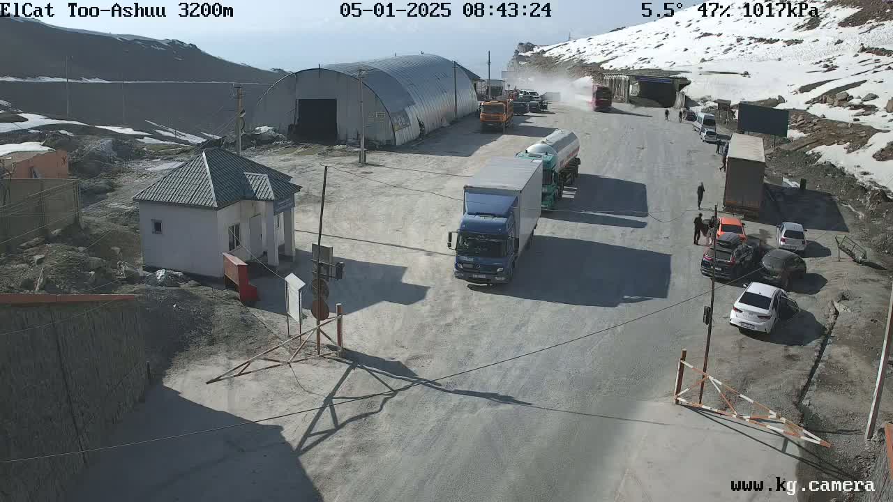 A mountain road checkpoint with several trucks and cars, a small building, and snowy mountains in the background under sunny conditions.