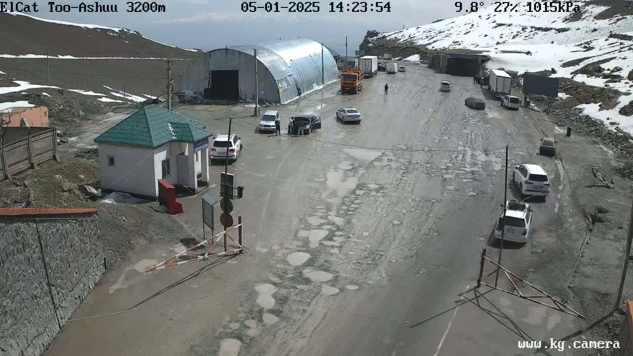 A muddy, pothole-ridden road at a mountain pass checkpoint under a partly sunny sky, with several vehicles and a small building present.