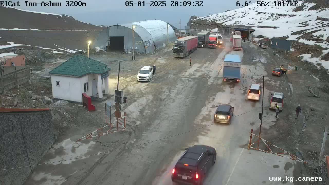 A mountain road with several vehicles, including trucks and cars, near a small building and a tunnel; it is overcast.