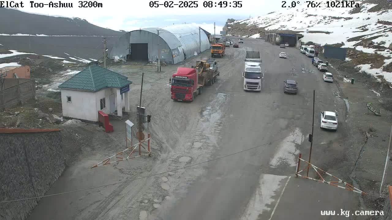 A mountain road with several vehicles, including trucks and cars, is partially covered in snow and has a small building near a gate.