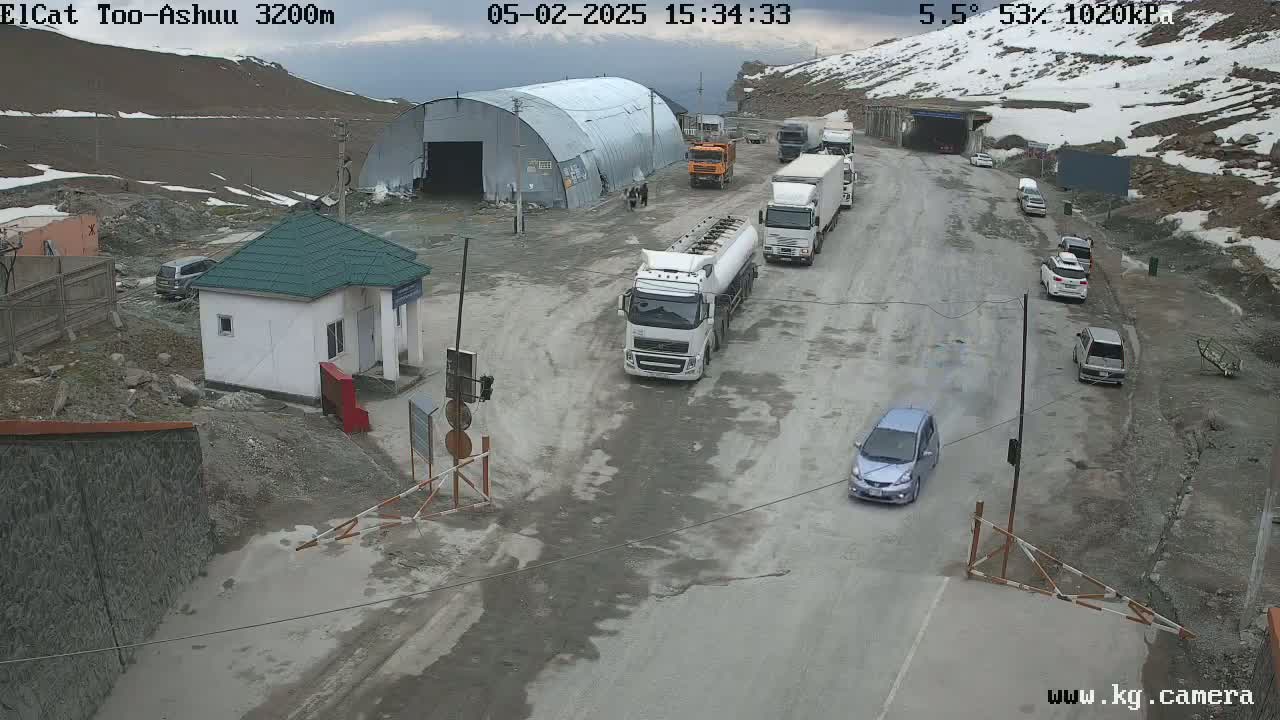 A mountain road with several vehicles, including trucks and cars, is seen near a small building and a large metal structure, with snow visible on the surrounding mountains under partly cloudy skies.