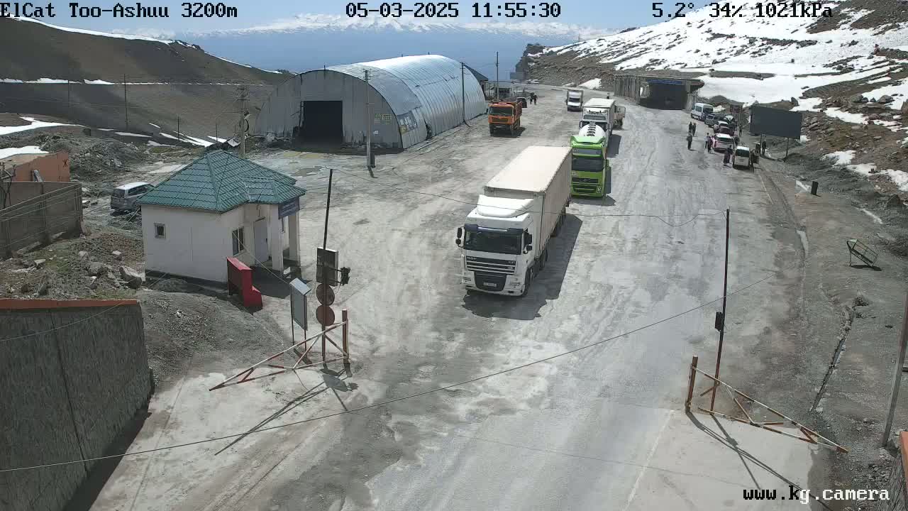 A mountain road scene on a sunny day shows several large trucks and a few cars parked near a small building, with snow visible on the surrounding mountains.