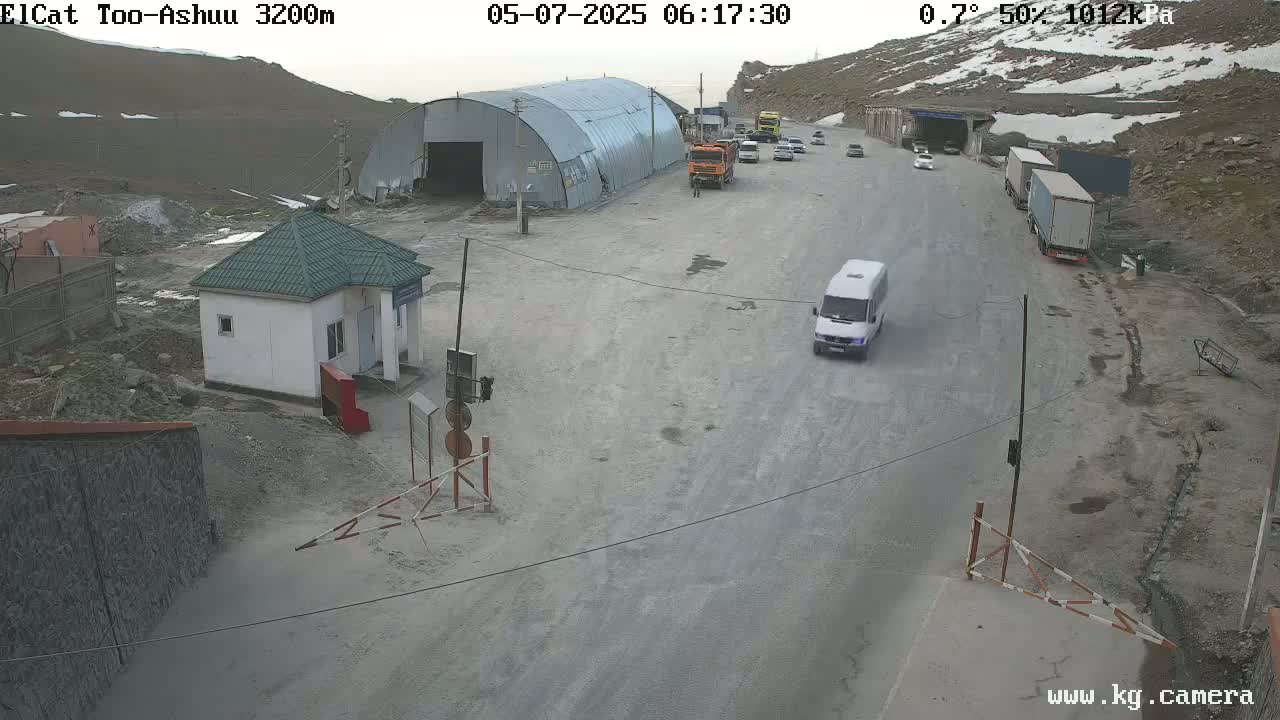 A mountain road with several vehicles, including a van and trucks, near a small building and a large metal structure, under partly cloudy conditions with some snow visible on the surrounding mountains.