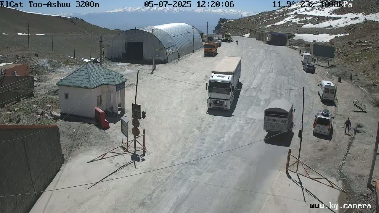 A mountain road on a sunny day with several vehicles parked near a small building and a large metal structure, with snow visible in the distance.