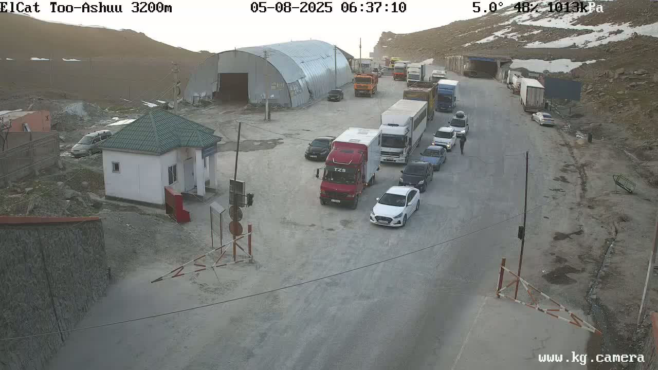 A line of vehicles waits at a mountain pass border crossing under partly cloudy skies with patches of snow visible on the surrounding hills.