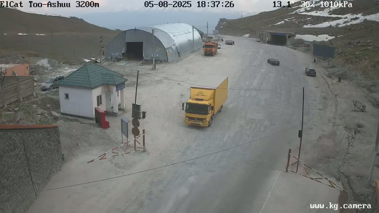 A mountain road with several vehicles, a small building, and a large metal structure, under partly cloudy skies.